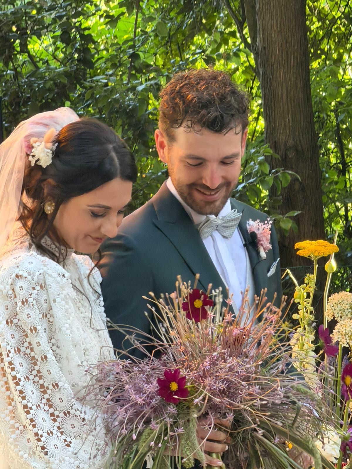 Couple holding a colorful bouquet outdoors under green trees, smiling and looking down together