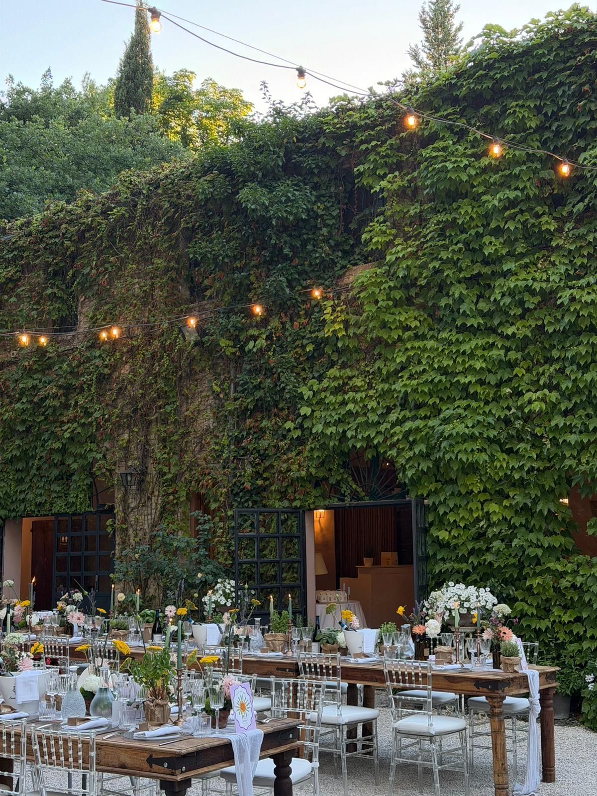 Outdoor dining setup under string lights beside a vine-covered wall with open doors and tables set for a reception