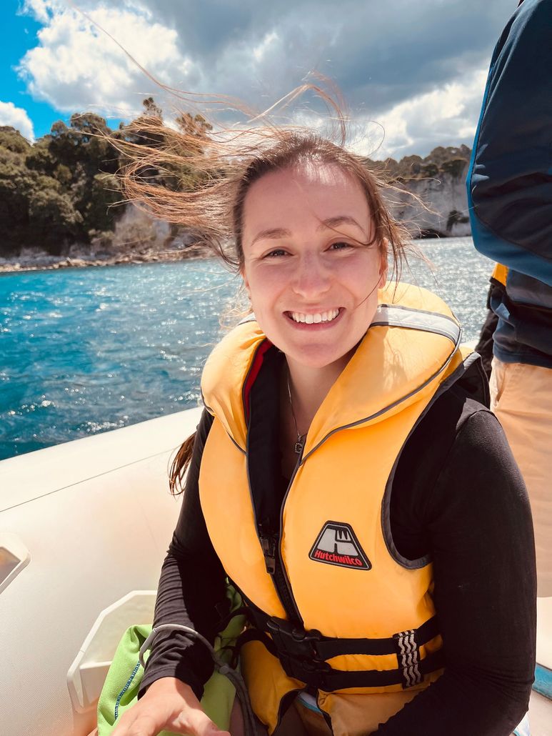 Smiling person in a yellow life jacket on a boat with turquoise water and rocky shoreline in the background
