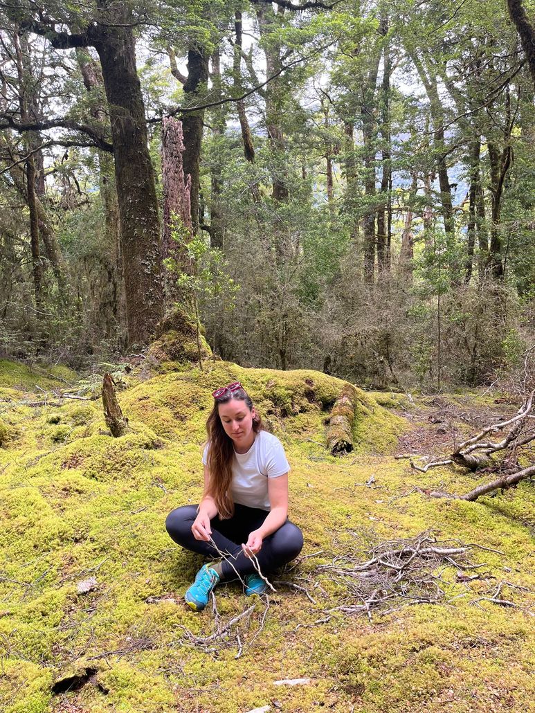 Person crouching on moss in a wooded forest, wearing a white shirt, black pants, and blue shoes.