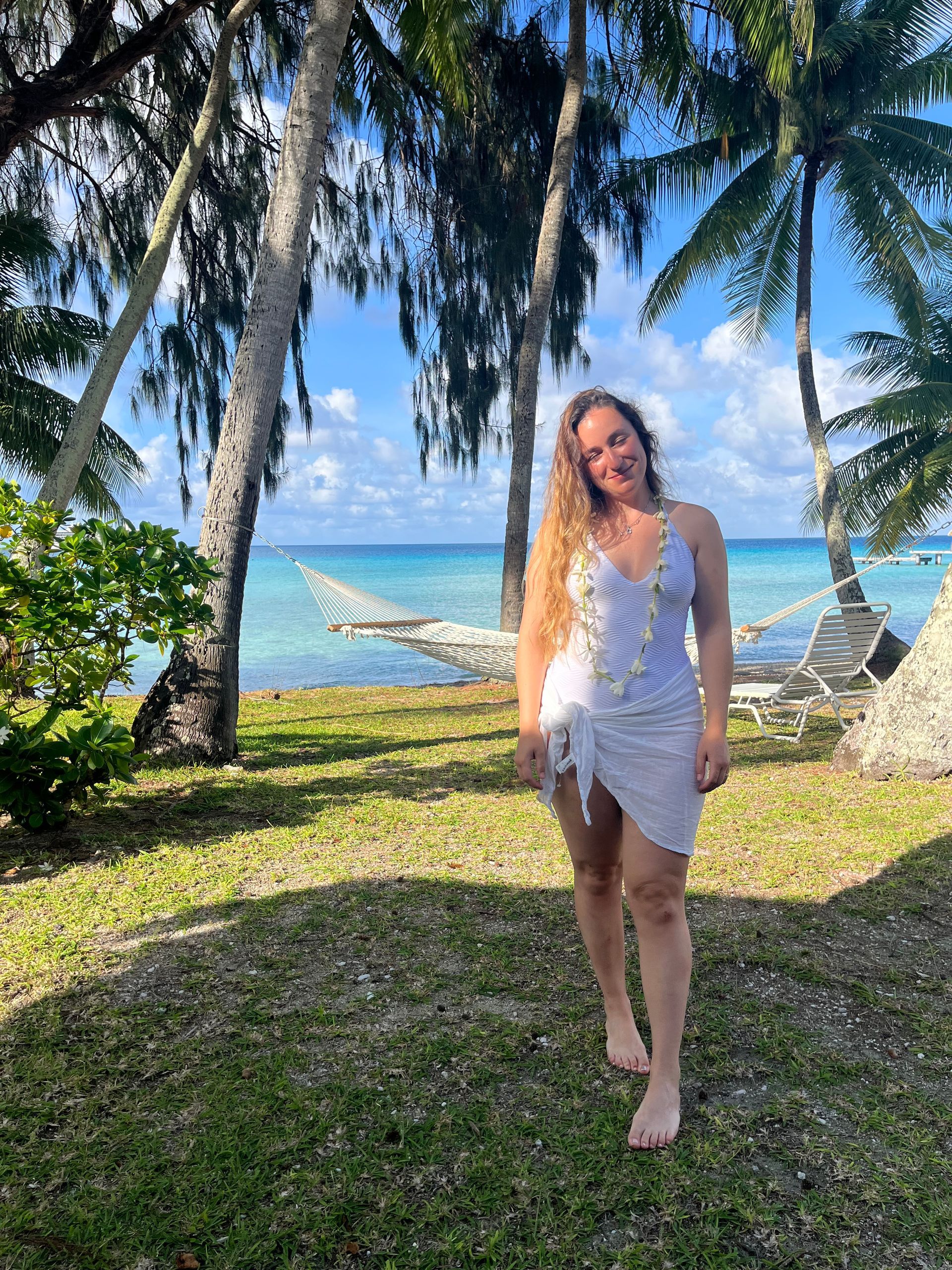 Woman in a white dress standing on a grassy beach with palm trees and ocean in the background
