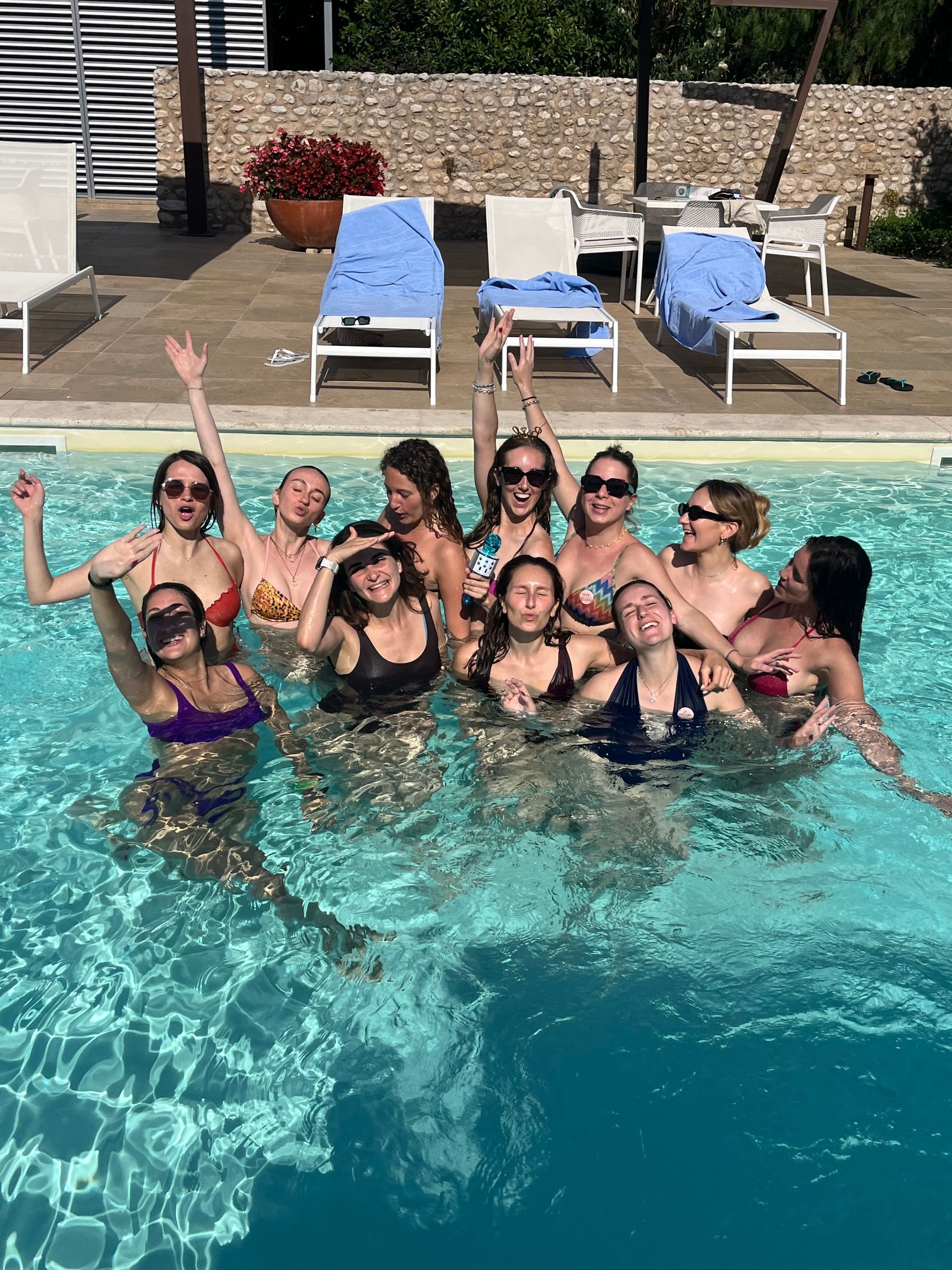Group of people smiling and posing in a swimming pool with raised arms, lounge chairs in the background