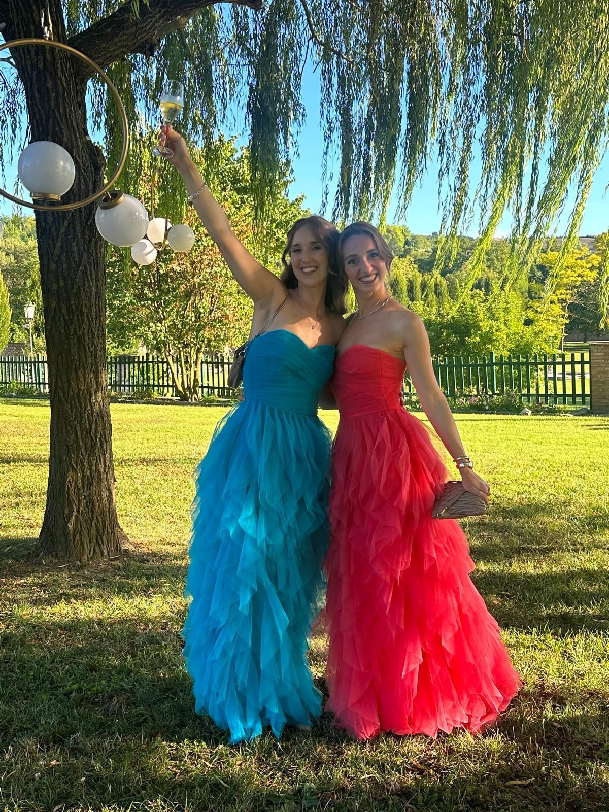 Two people in bright blue and red dresses posing outdoors under a tree, smiling and raising their arms