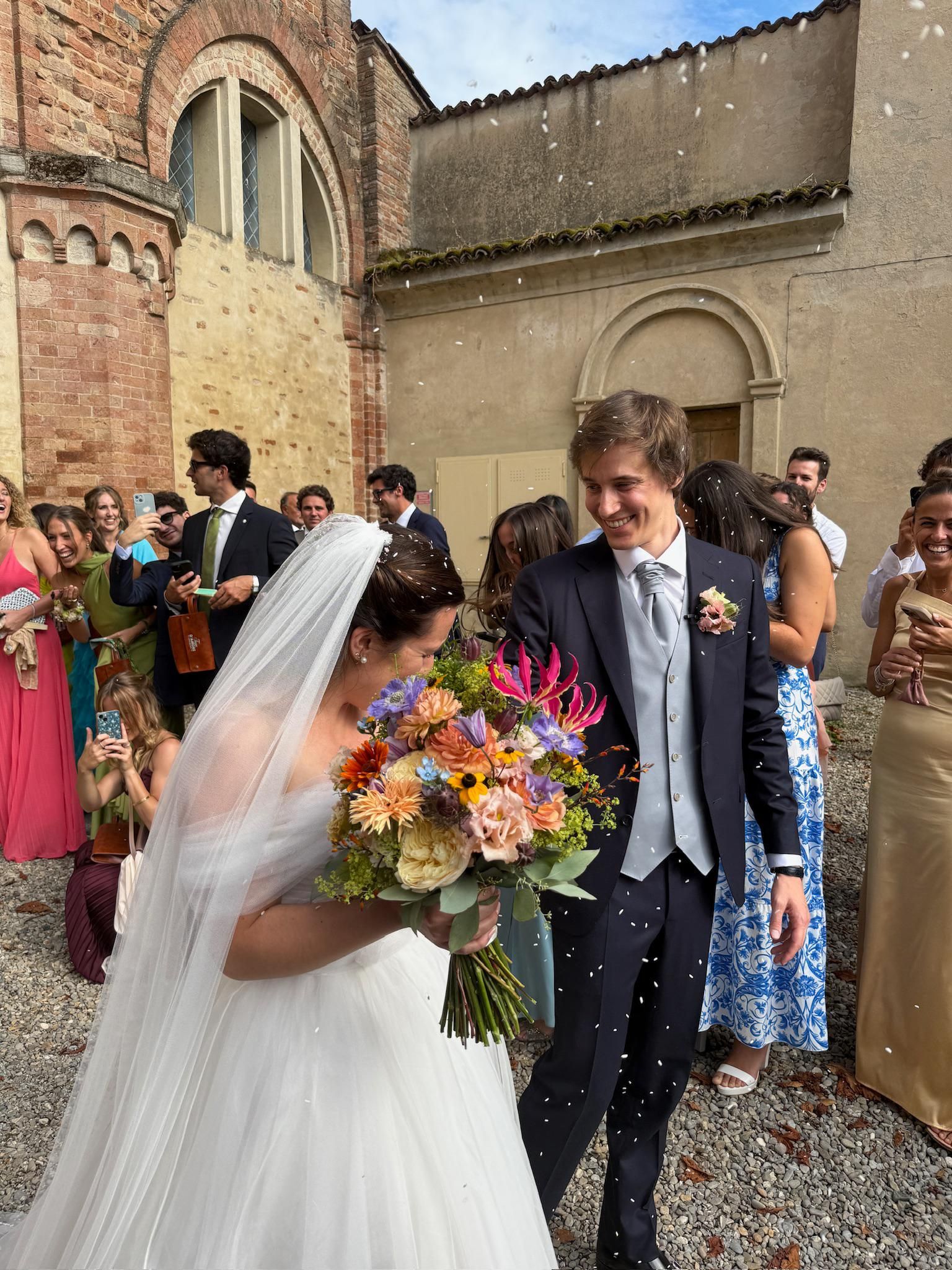 Wedding couple walking through confetti outside a church, bride holding a colorful bouquet