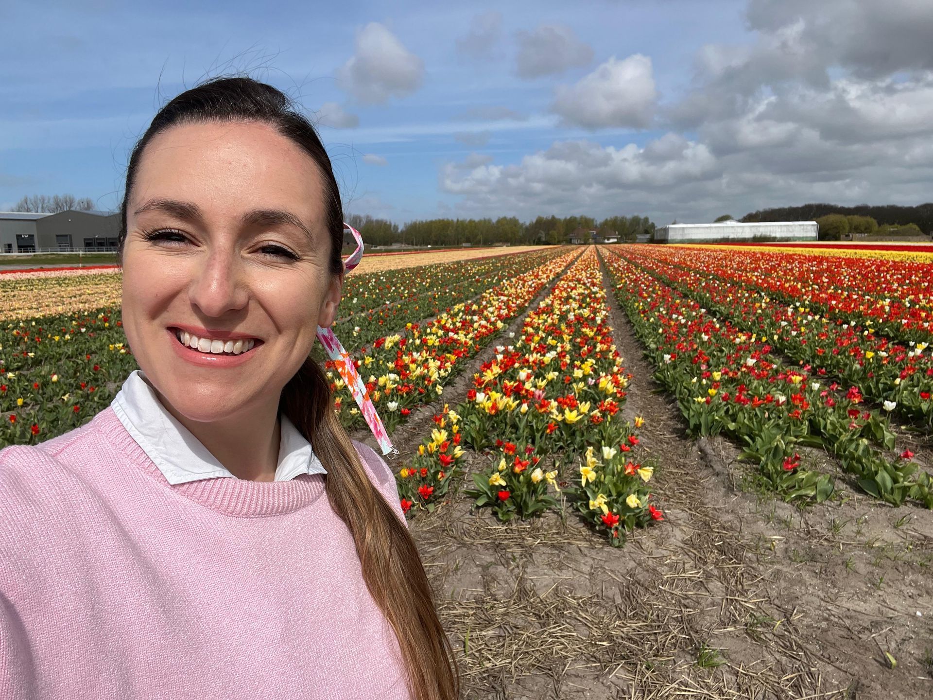 Selfie of a smiling woman in a pink sweater in front of colorful tulip rows under a blue sky.