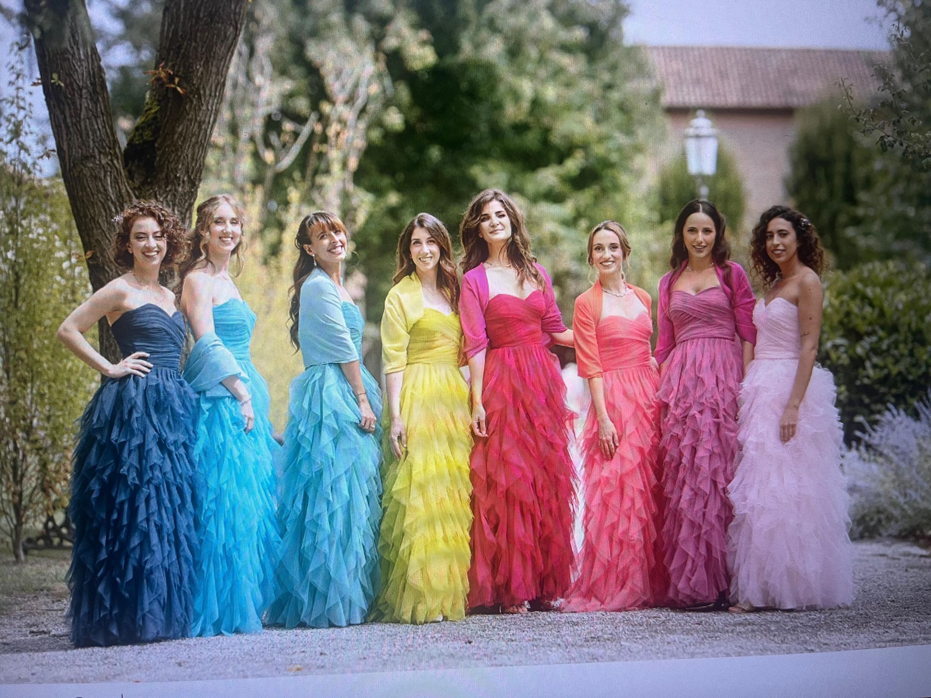 Seven women in bright ruffled gowns standing outdoors in a row, posing together in a garden.