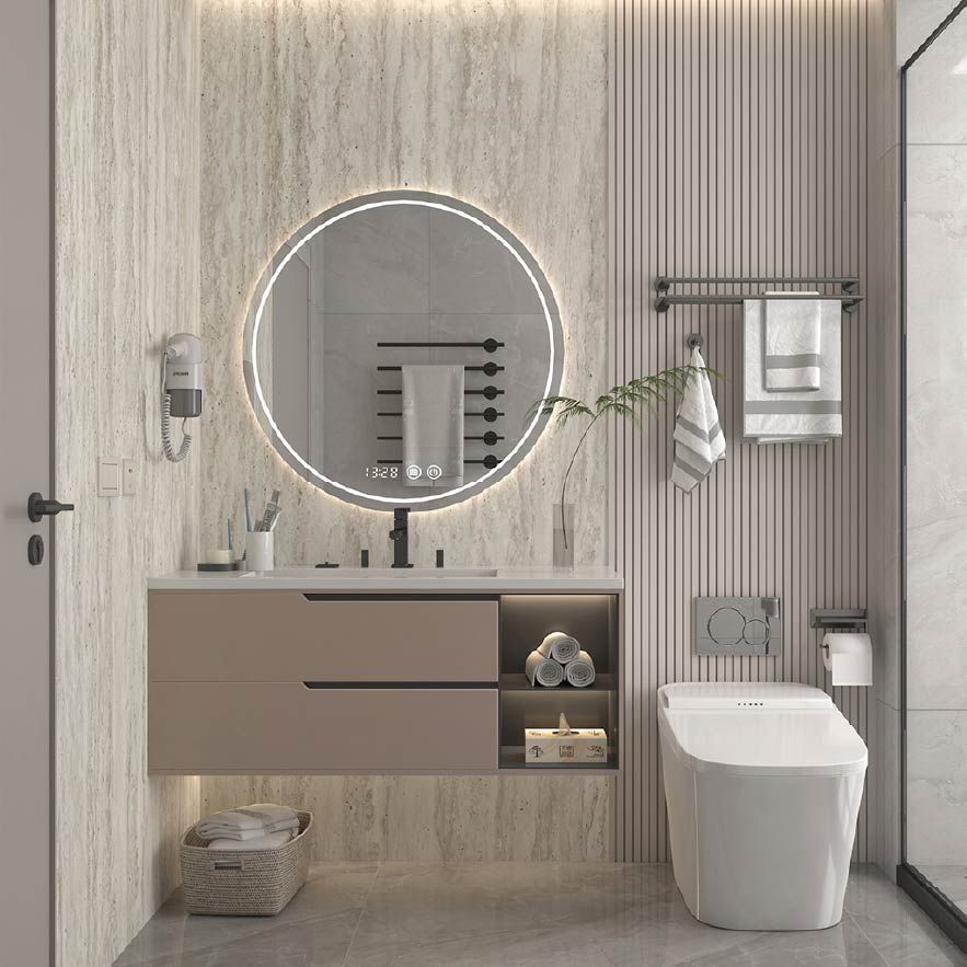 Baño moderno de color beige con espejo redondo iluminado, lavabo suspendido, inodoro, toallero y panel de vidrio para la ducha.