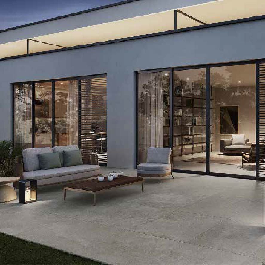 Patio moderno con sofá exterior gris, tumbona y puertas correderas de cristal que dan a un interior con iluminación cálida.