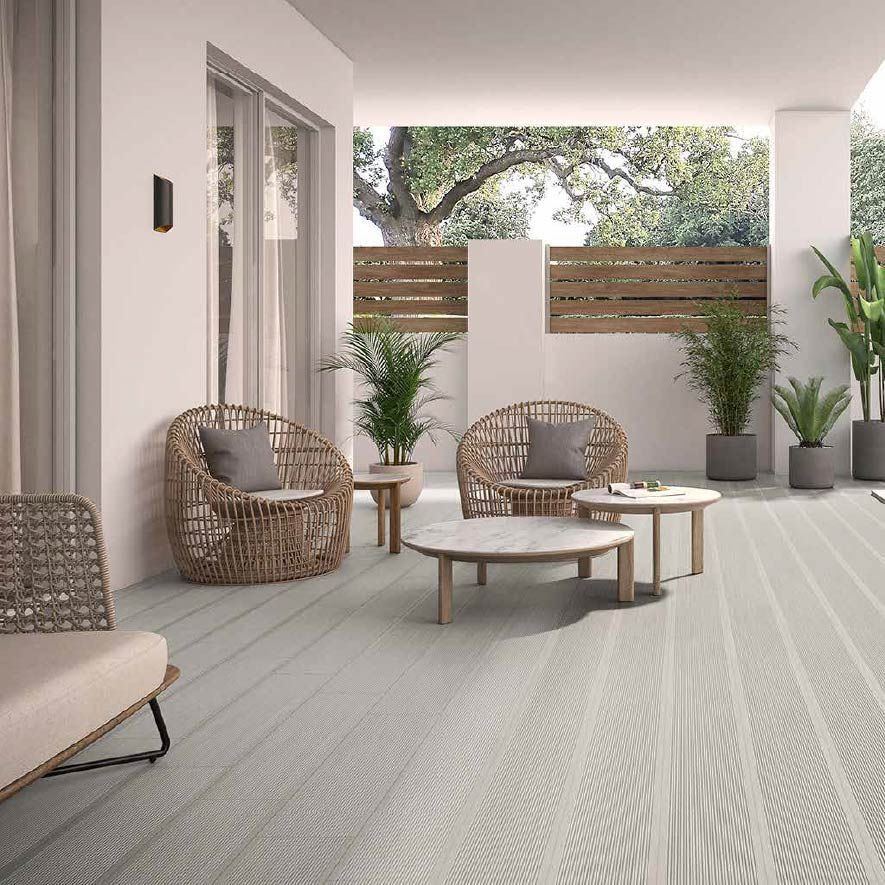 Patio moderno con sillas de mimbre, mesas bajas, plantas en macetas y una alfombra gris claro bajo un porche cubierto.