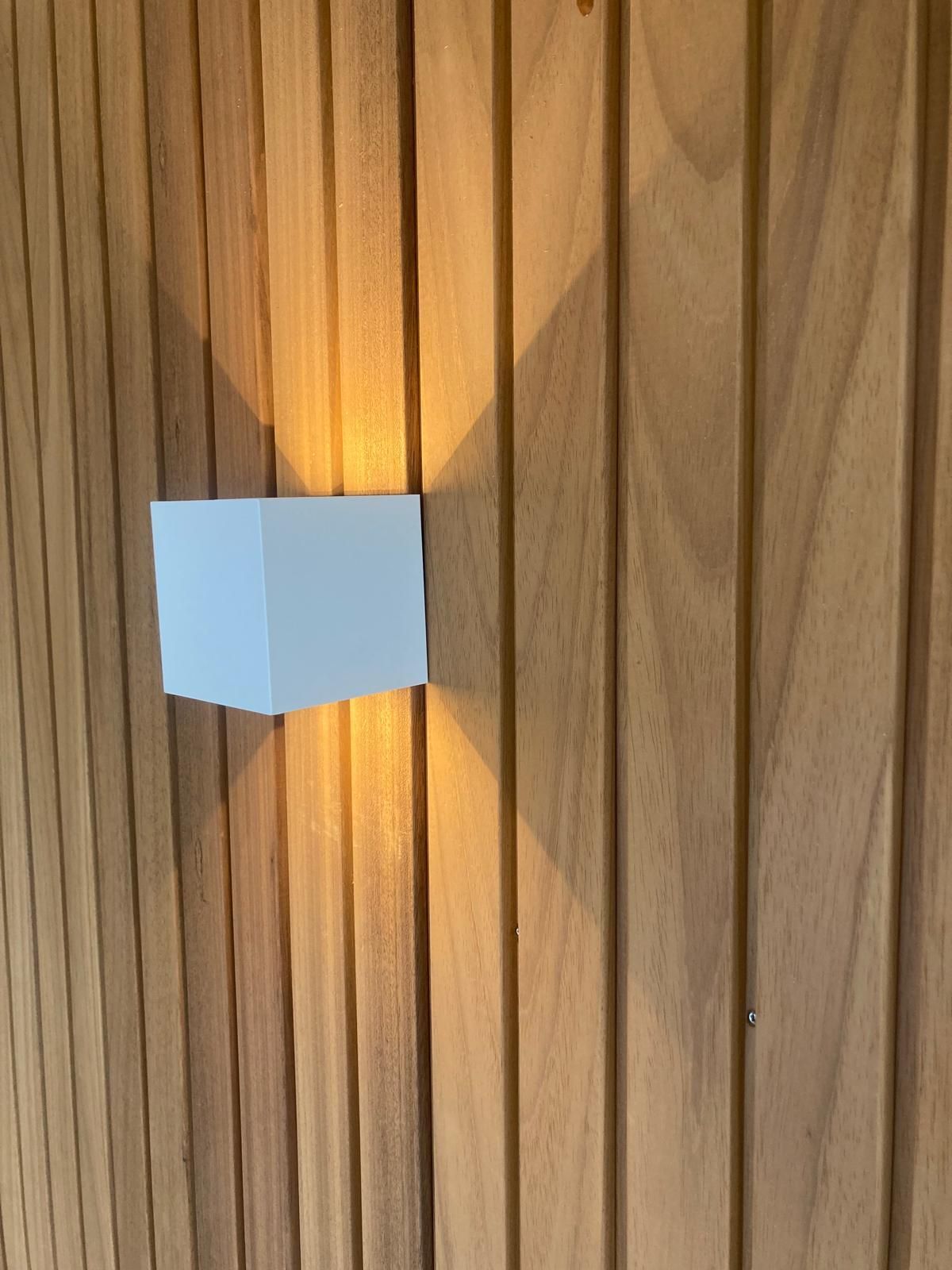 Een moderne, witte, kubusvormige wandlamp verspreidt een warm licht tegen een wand bekleed met verticale houten latten.