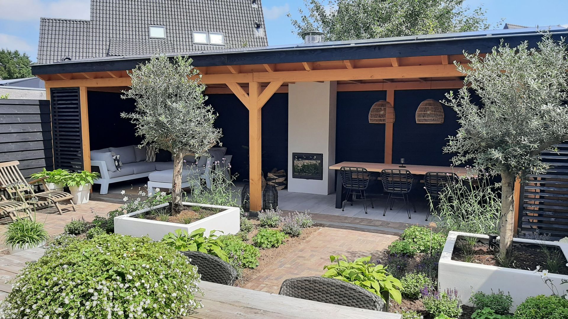 Een overdekt terras met een open haard, een eethoek en comfortabele zitgelegenheid, geflankeerd door twee olijfbomen in witte plantenbakken.