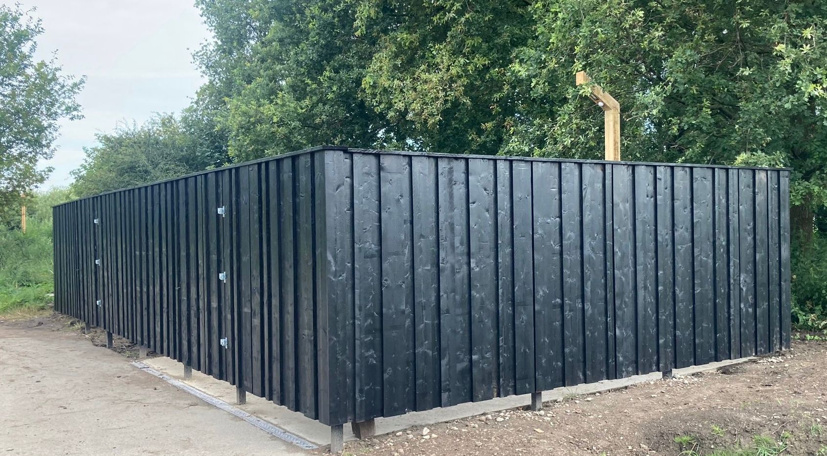 Een moderne, zwartgeverfde verticale schutting van houten latten staat buiten op een grindterrein met weelderige groene bomen op de achtergrond.