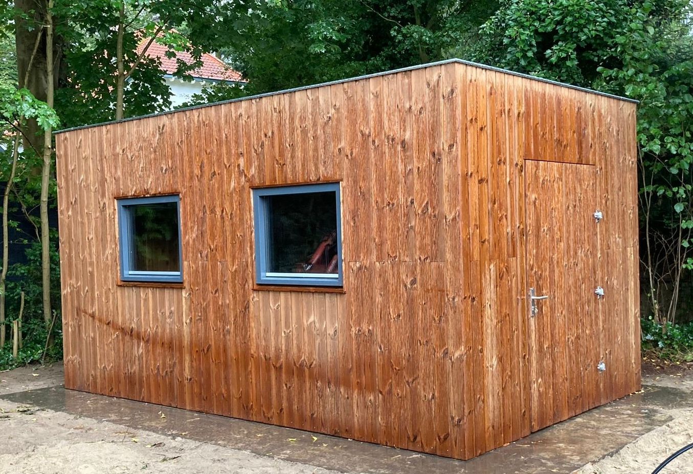 Een rechthoekig houten schuurtje met twee ramen en een deur, gelegen in een achtertuin omgeven door bomen.