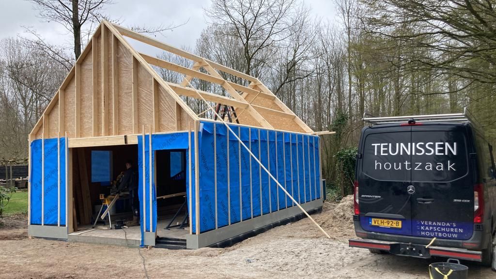 De constructie van een houten schuurframe, omwikkeld met blauwe beschermfolie, naast een servicebus met bedrijfslogo in een bosrijk gebied.