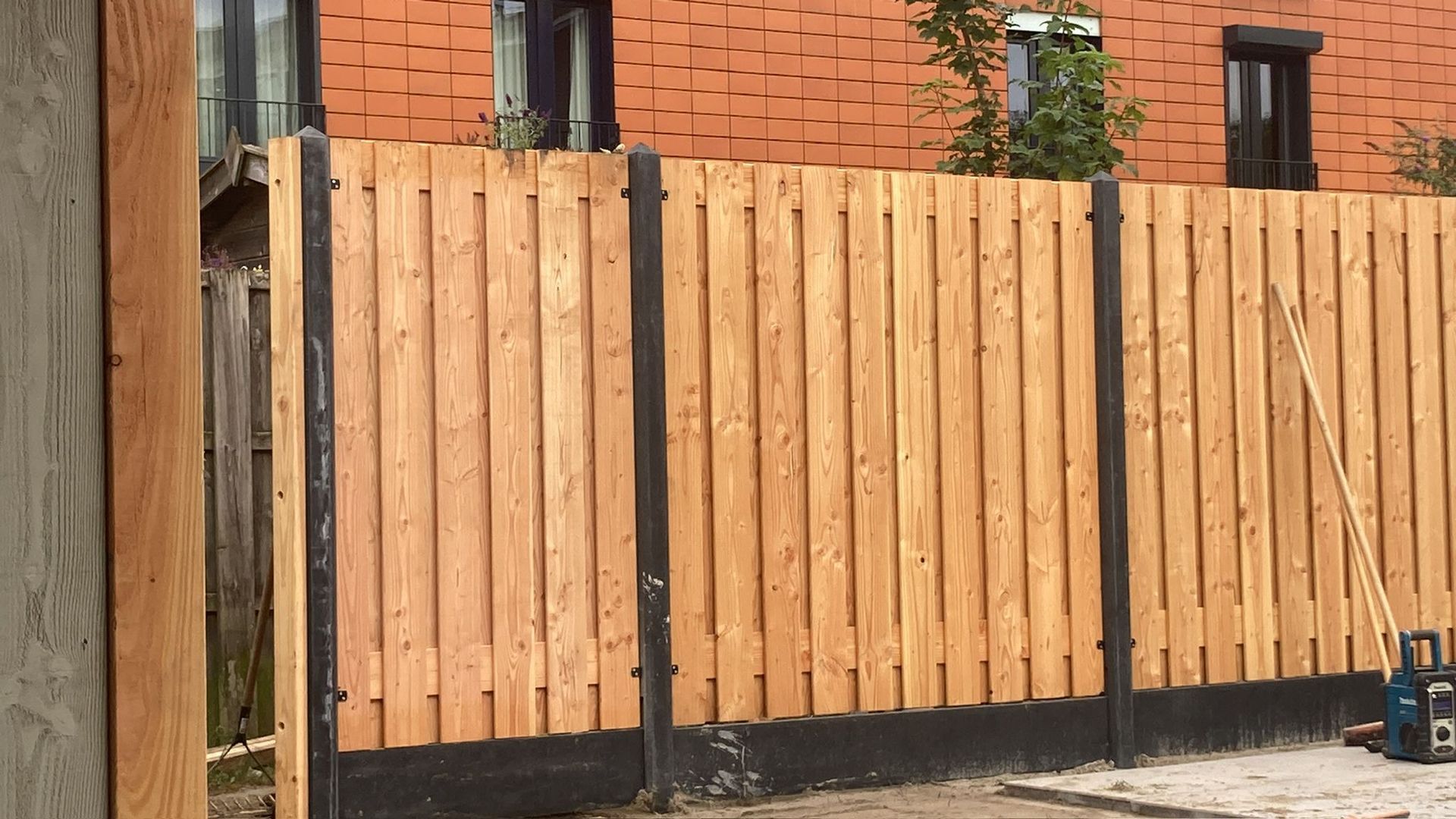 Voor een gebouw van rode baksteen staat een nieuwe houten schutting met verticale latten en donkere betonnen palen.