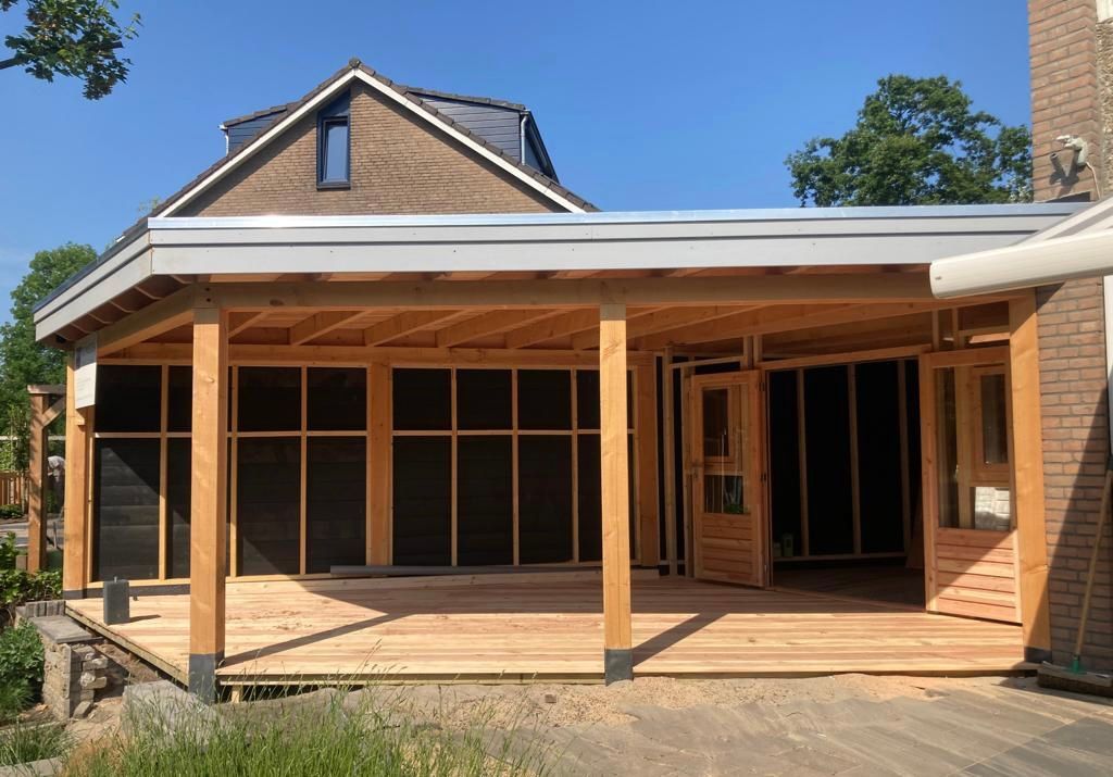 Een houten terrasuitbouw met een groot dak, aangebouwd aan een bakstenen huis, voorzien van glazen deuren en een houten vlonder.