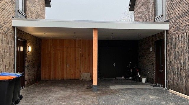 Een carport tussen twee bakstenen huizen, met een houten muur aan de linkerkant, een donkere deur aan de rechterkant en een centrale pilaar.
