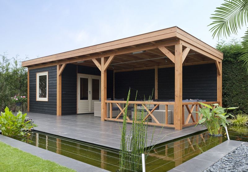 Een moderne zwarte schuur met een overdekte houten veranda en tuinmeubilair, gelegen naast een aangelegde tuinvijver.