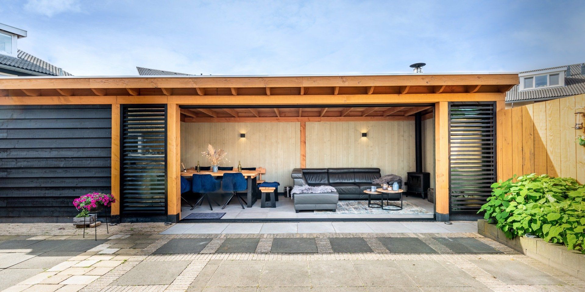 Een openlucht houten buitenruimte met een eethoek en een loungegedeelte, voorzien van een grijze bank en donkere lamellenluiken.