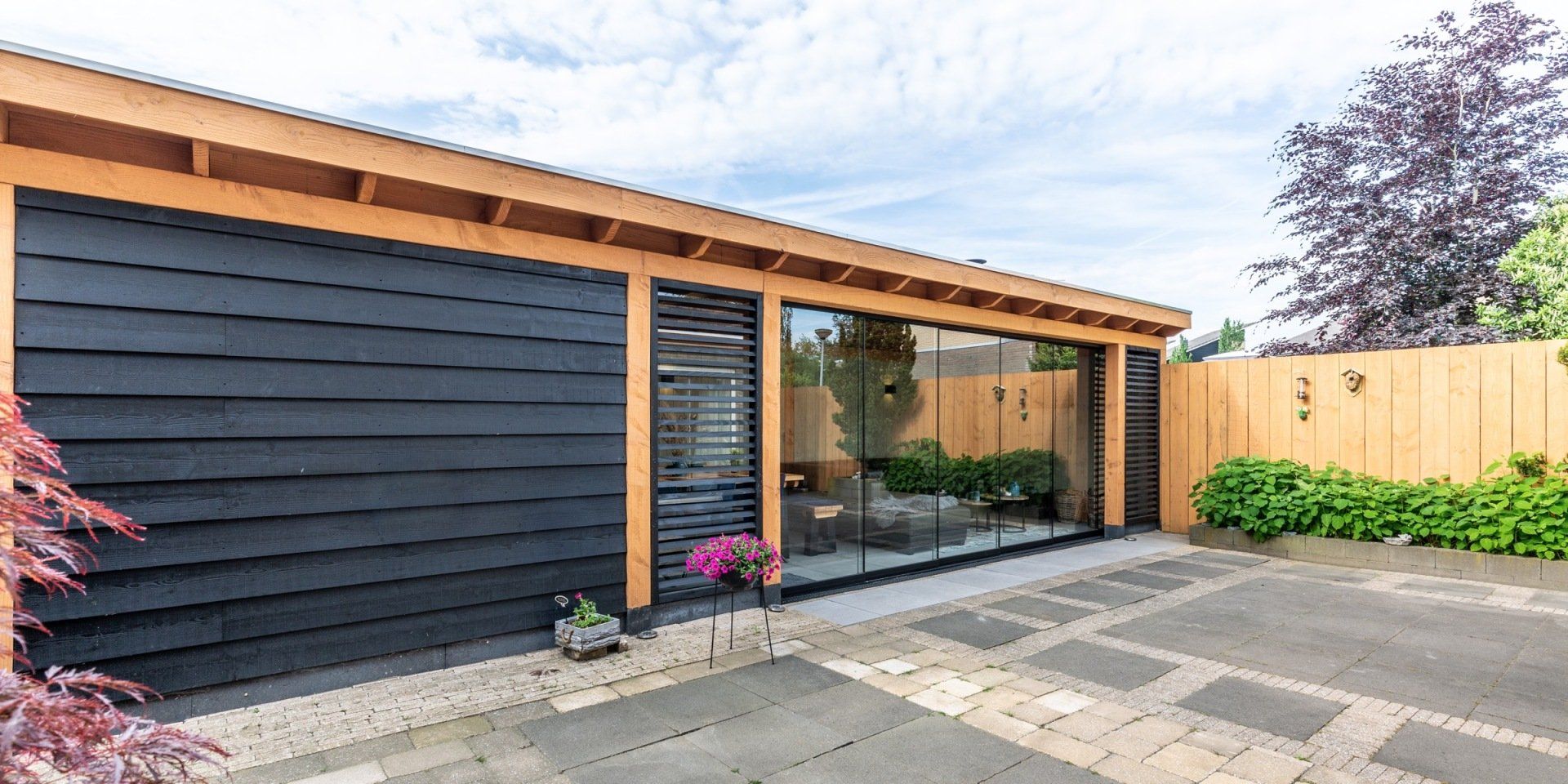 Moderne tuinkamer met zwarte houten gevelbekleding, glazen wanden, een houten frame en een betegeld terras, gezien op een zonnige dag.