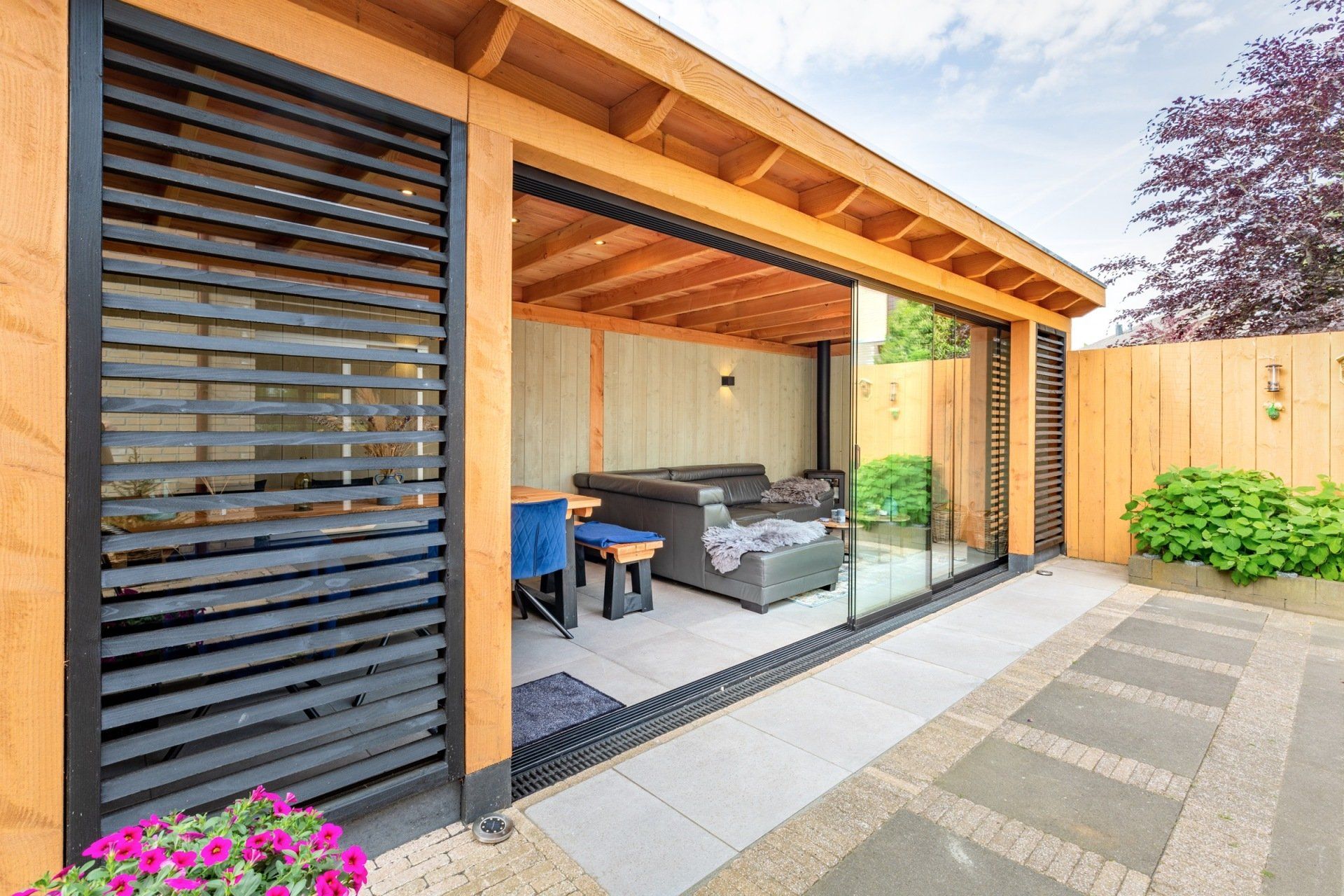 Een modern houten terraspaviljoen met schuifbare glazen wanden, lamellenluiken en een buitenlounge op een stenen terras.