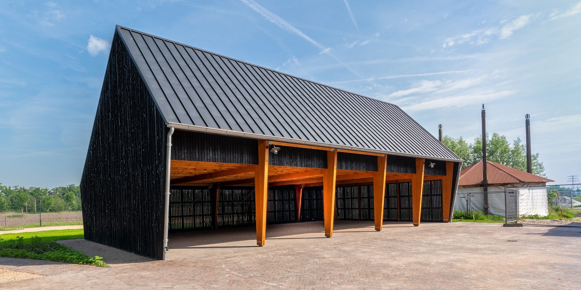 Een modern, openlucht paviljoen van hout met zwarte houten shingles en een zadeldak staat op een grindterrein onder een blauwe hemel.