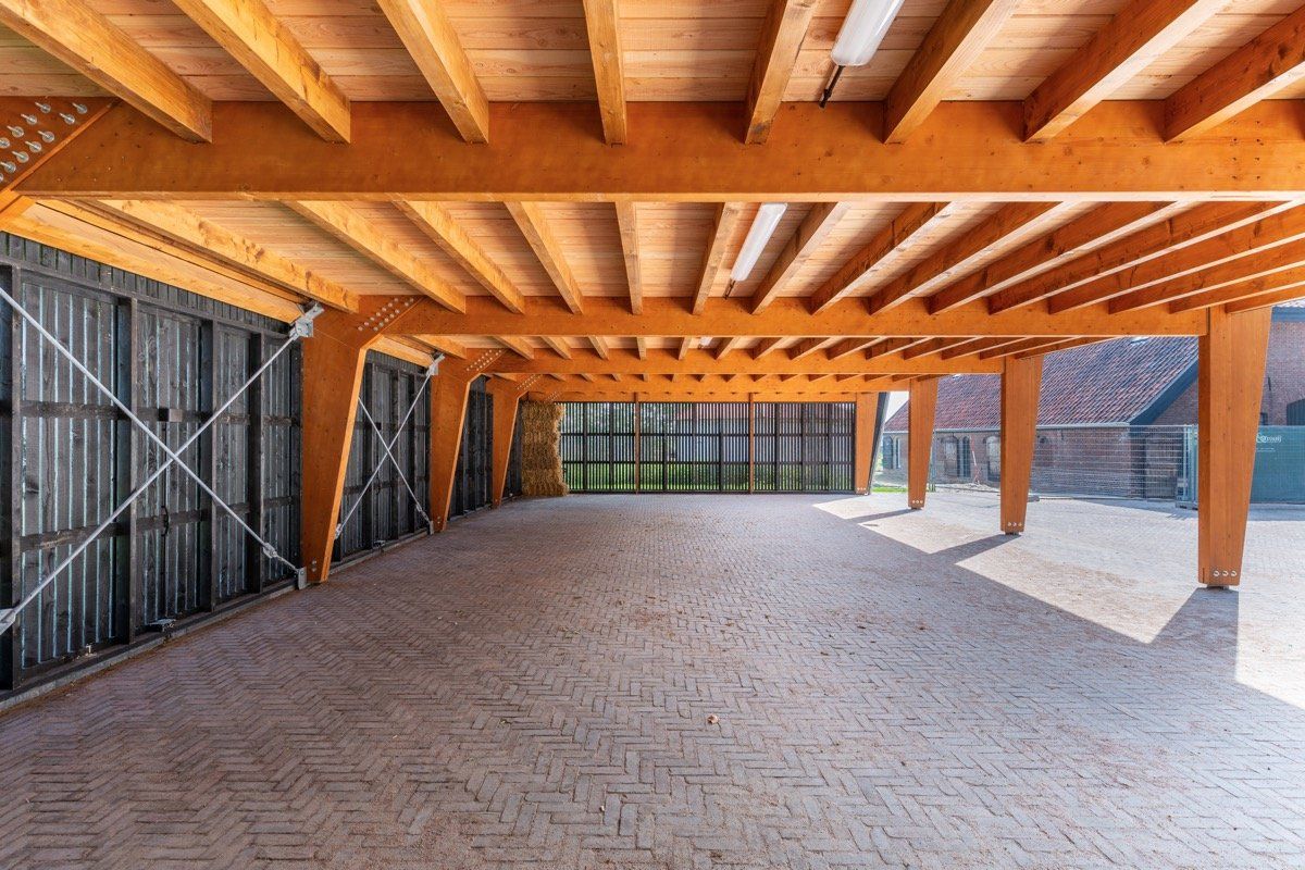 Een open houten carport met een bestrate vloer, voorzien van zichtbare balken en donkere lattenwanden onder een overdekt plafond.