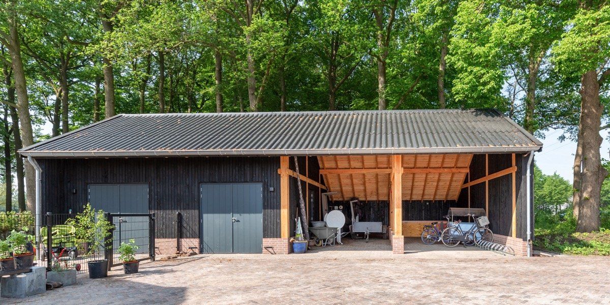 Een zwarte houten schuur met een pannendak, gesloten grijze deuren en een open carport, staat tegen de achtergrond van een bos.