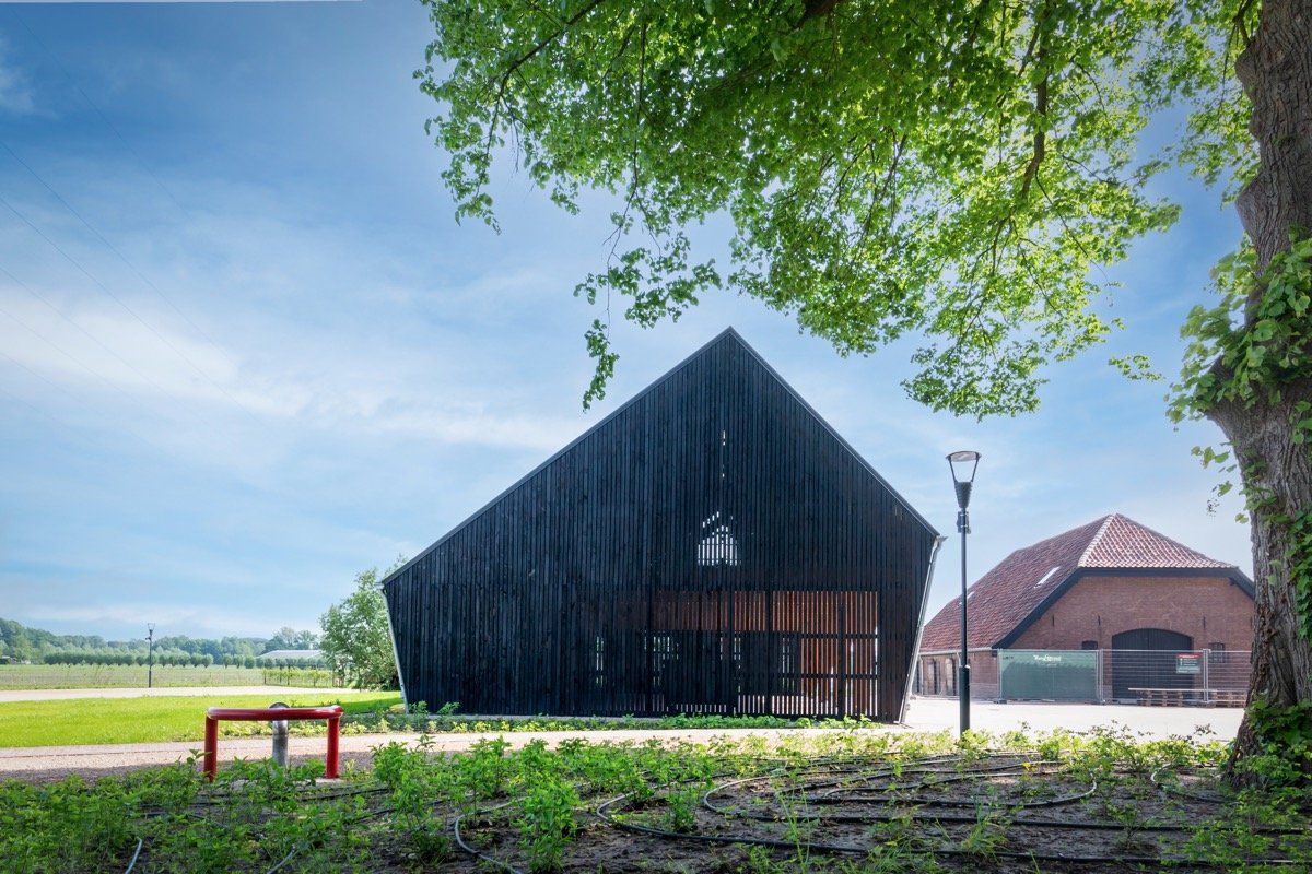 Een zwart, hoekig, modern gebouw in schuurstijl staat in een groen landschap naast een traditionele boerderij van rode baksteen.