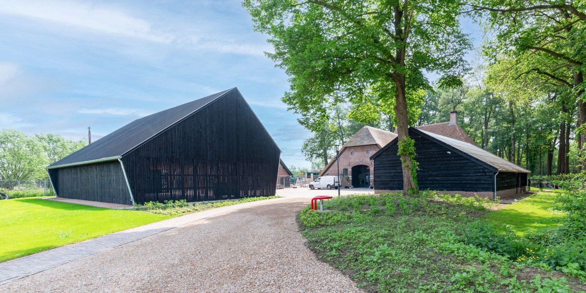 Een grindpad leidt naar twee zwarte, houten schuurachtige gebouwen op een grasveld omgeven door bomen.