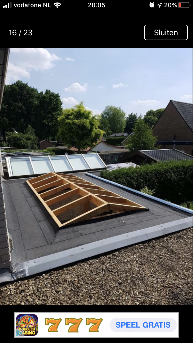 Houten frame voor een dakraam dat wordt geïnstalleerd in een plat gebouw met een zwart dak, met op de achtergrond bomen en huizen.