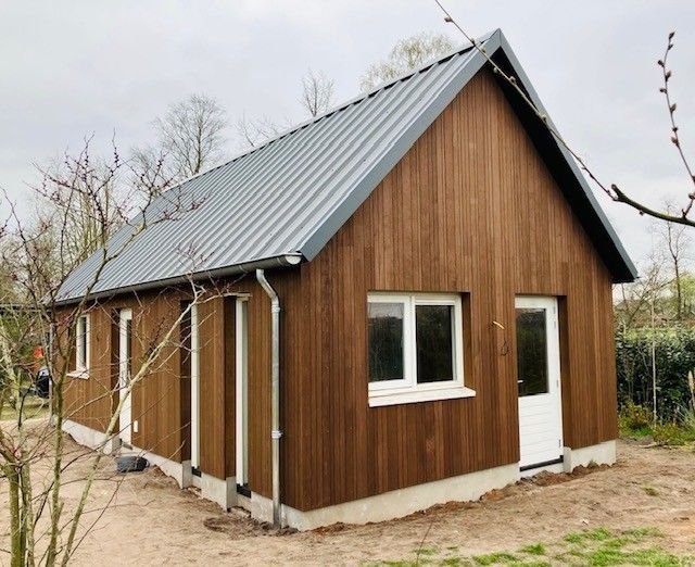 Een moderne, gelijkvloerse blokhut met bruine verticale houten gevelbekleding, een grijs metalen dak en witte deuren en ramen.