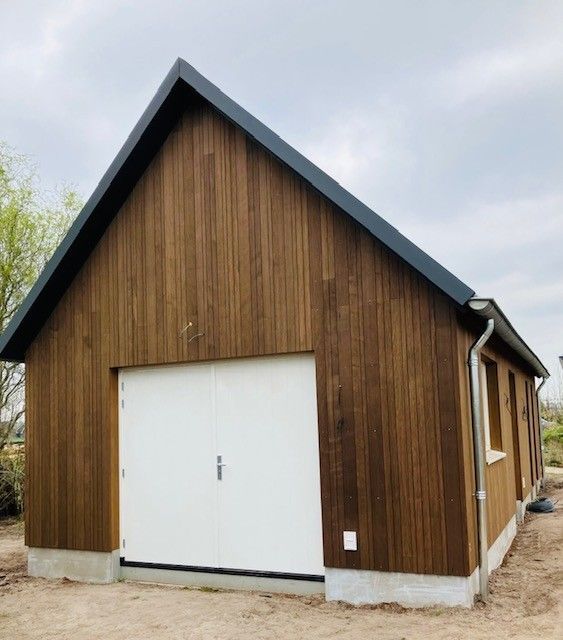 Een vrijstaande houten garage met verticale gevelbekleding, een witte dubbele deur en een donker metalen zadeldak onder een bewolkte hemel.