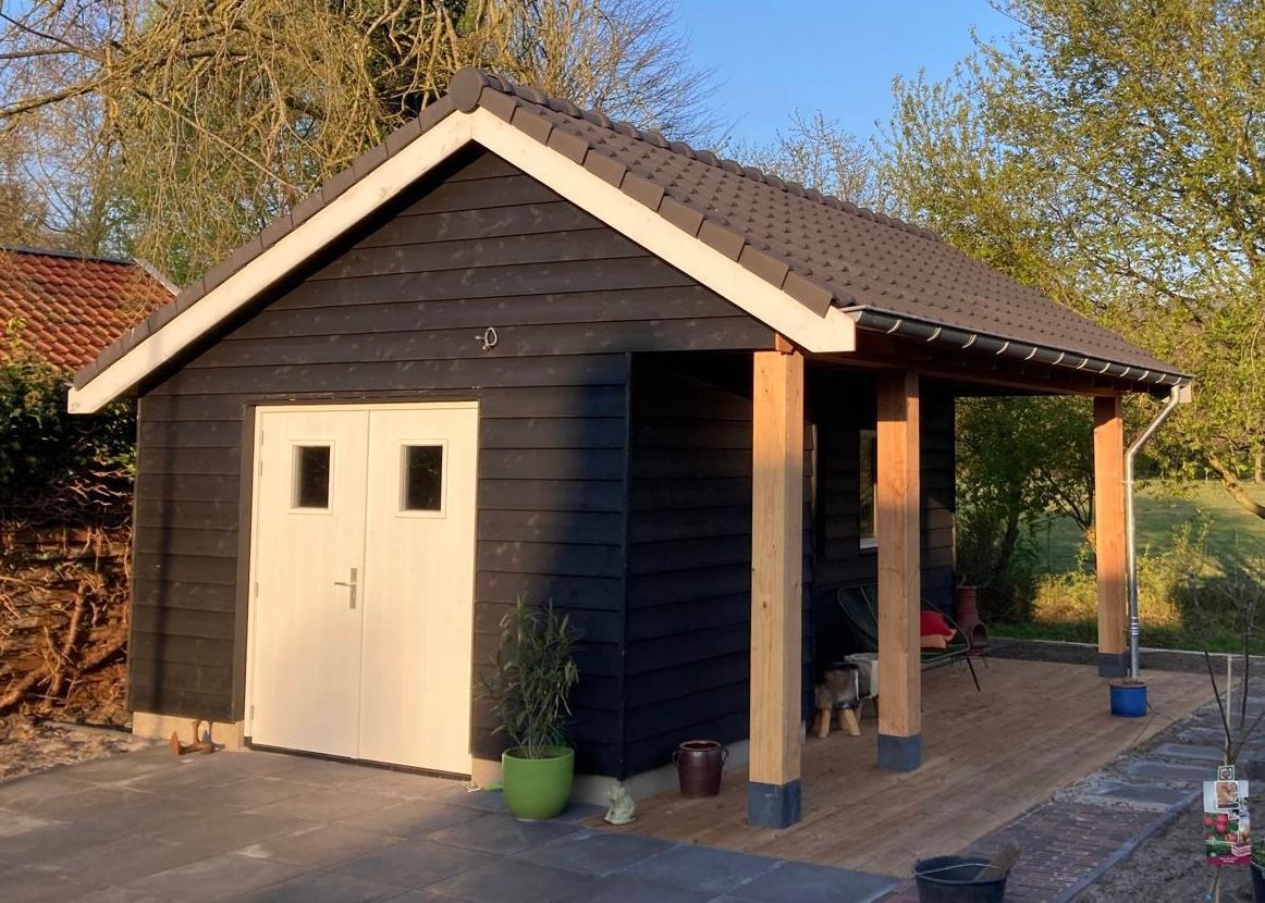 Een zwarte houten schuur met witte dubbele deuren, een pannendak en een open zijveranda met houten pilaren.