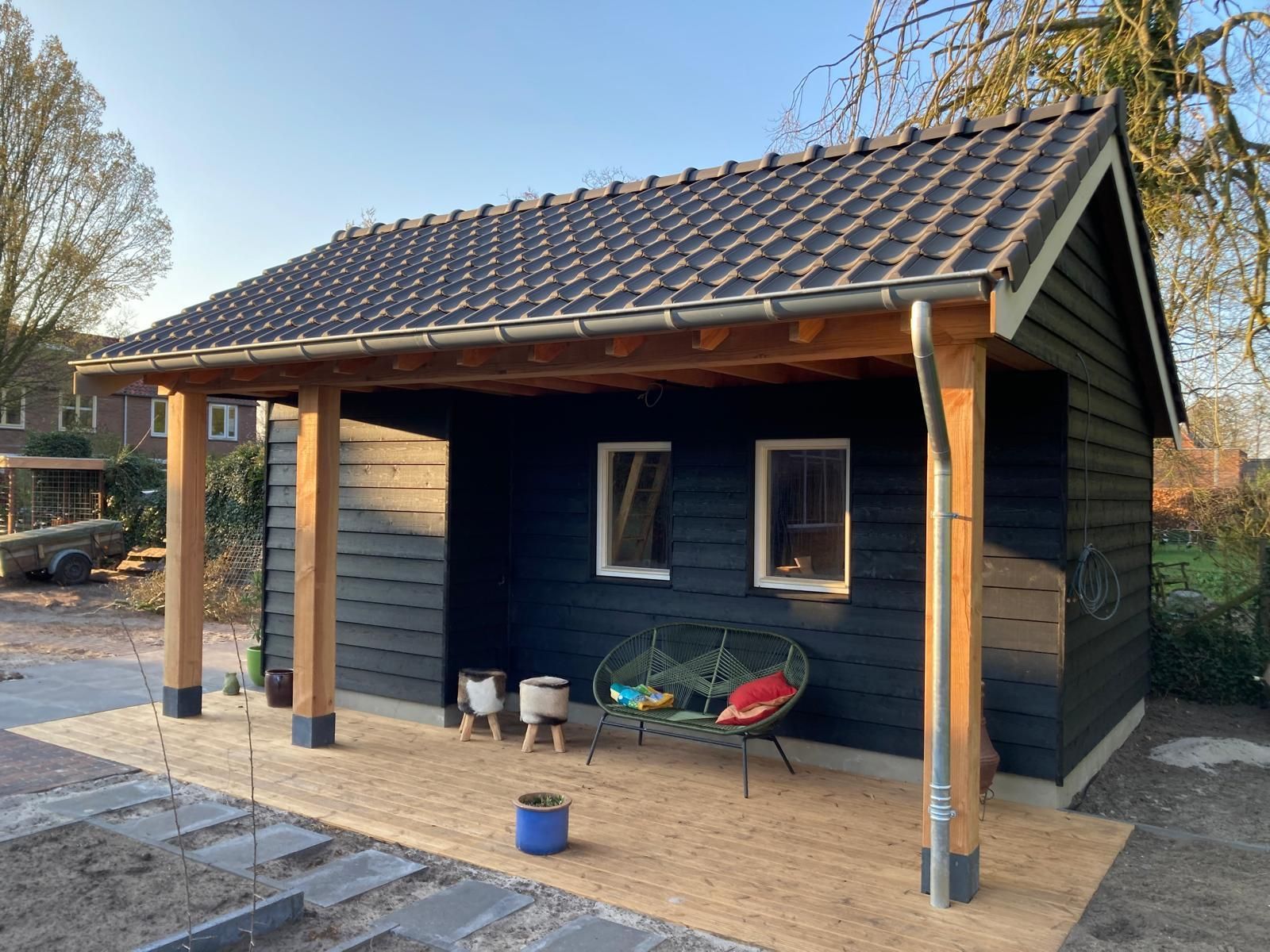 Een zwart houten tuinhuisje met een pannendak, een overdekte veranda, twee kleine ramen en een bankje op een houten terras.