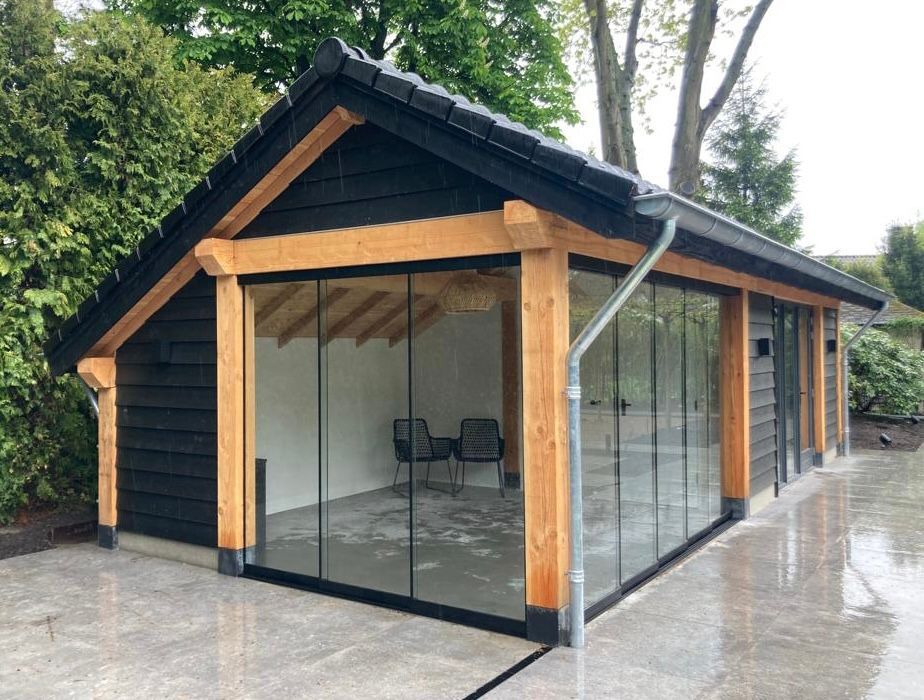 Een moderne buitenkamer voor in de tuin met zwarte houten gevelbekleding, een frame van natuurlijk hout en glazen schuifwanden op een stenen terras.