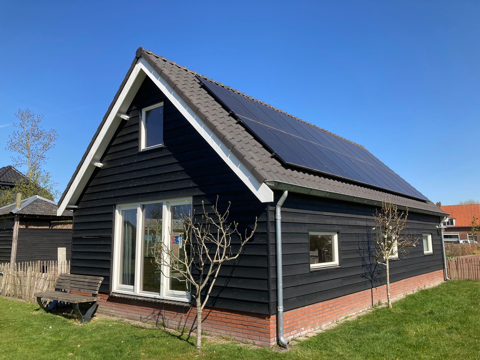 Een moderne, zwarte houten blokhut met een zadeldak, zonnepanelen en een fundering van rode bakstenen, gelegen op een grasveld onder een blauwe hemel.