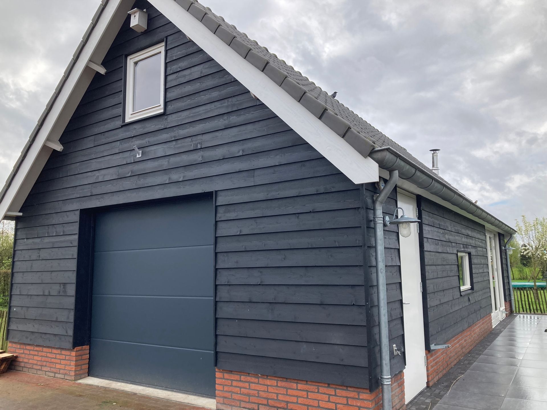 Een vrijstaande garage met donkergrijze horizontale gevelbekleding, een bakstenen fundering, een grijze sectionaaldeur en een klein zolderraam.