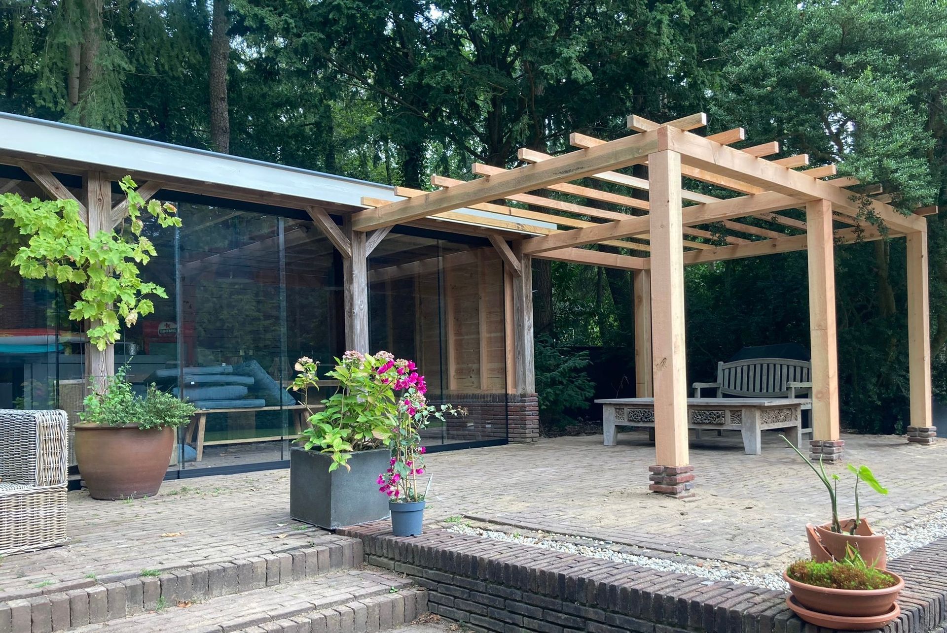 Een stenen terras met een moderne, glazen overkapping, een houten pergola, potplanten en tuinmeubilair.