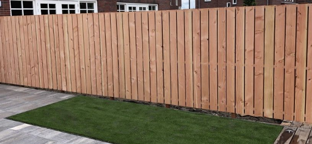 Een moderne houten schutting met verticale latten omzoomt een tuin met een klein stukje gras en een betegeld terras.