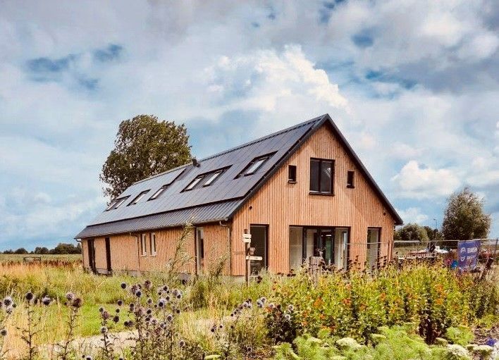 Een moderne woning in schuurstijl met verticale houten gevelbekleding en een donker metalen dak, gelegen in een veld met wilde bloemen onder een blauwe hemel.