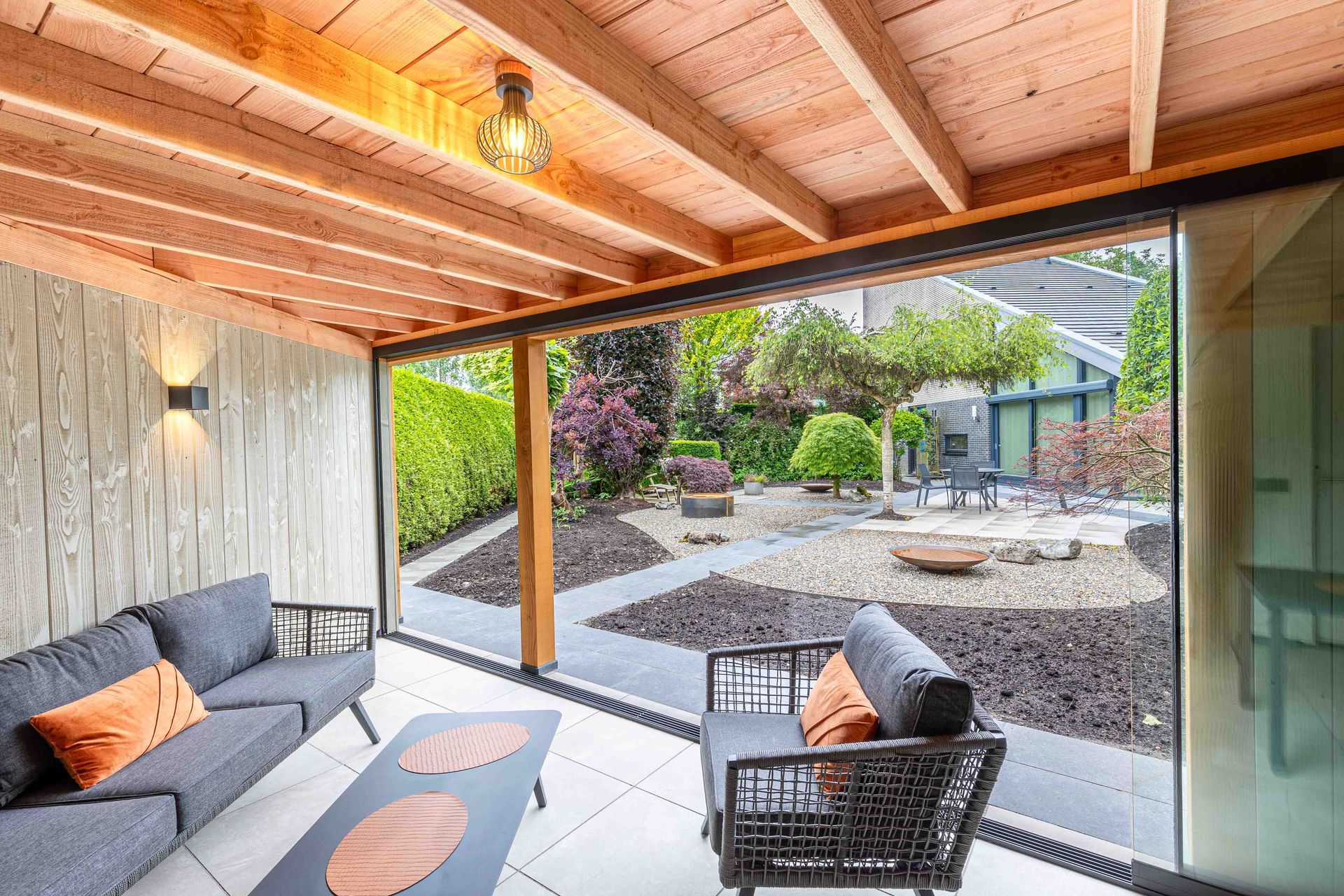 Moderne overdekte patio met donkergrijze tuinmeubelen, een tafel en een plafond met houten lambrisering, met uitzicht op een aangelegde tuin.