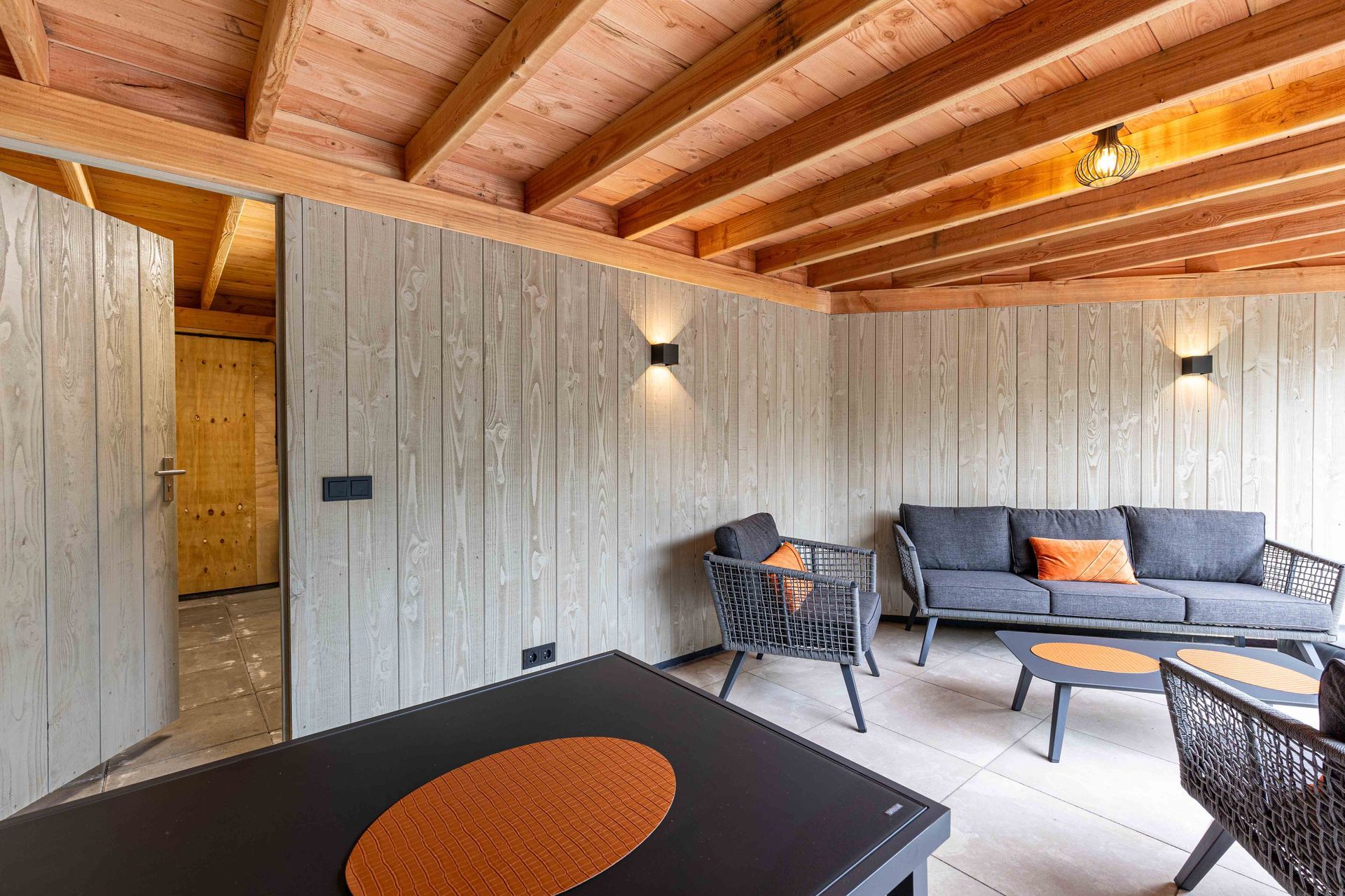 Een knusse kamer met zichtbare houten balken, lichtgrijze lambrisering, grijze meubels en een zwarte tafel met een oranje placemat.