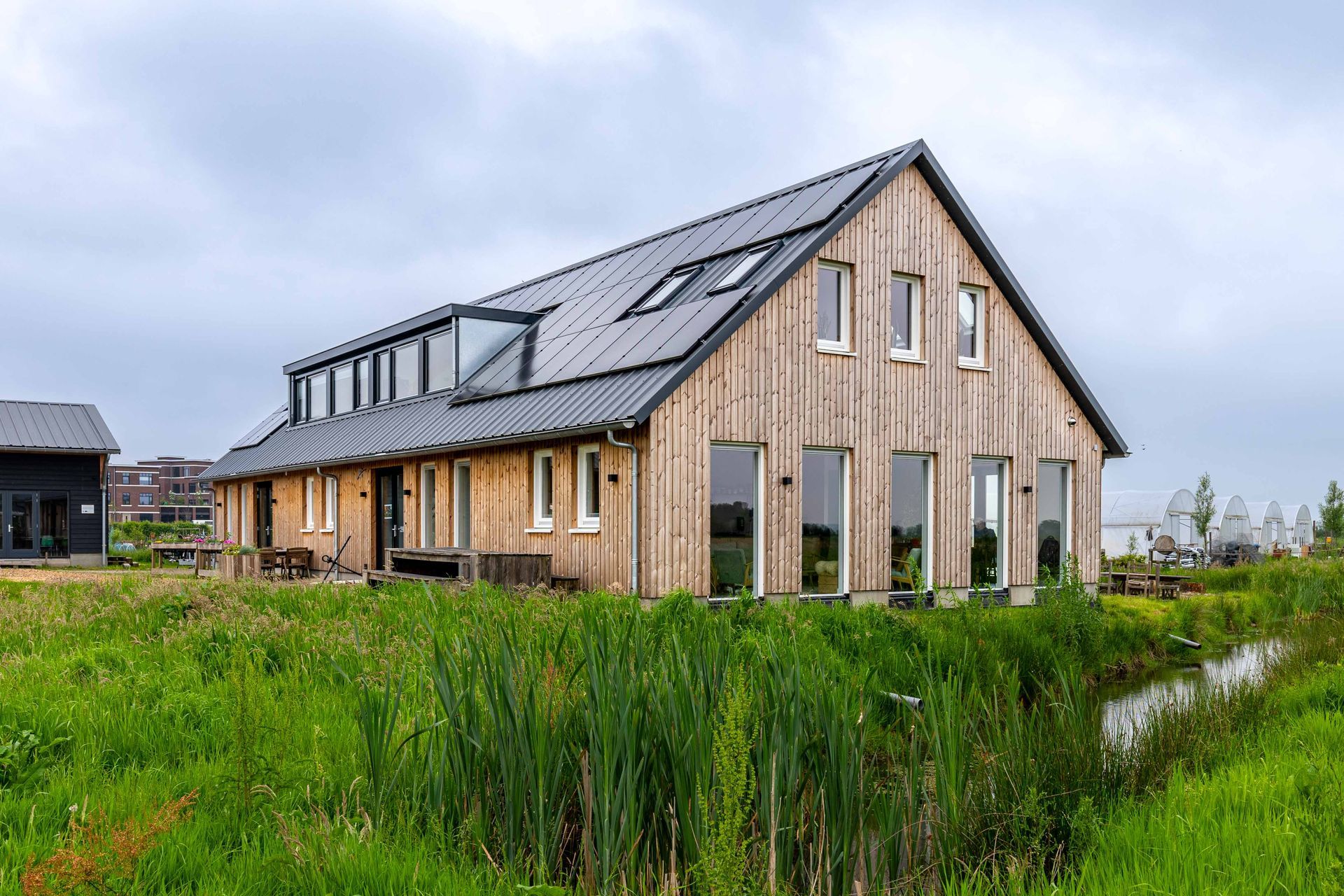 Moderne houten woning met een metalen dak en dakramen, gelegen naast een grasveld en een klein waterkanaal.