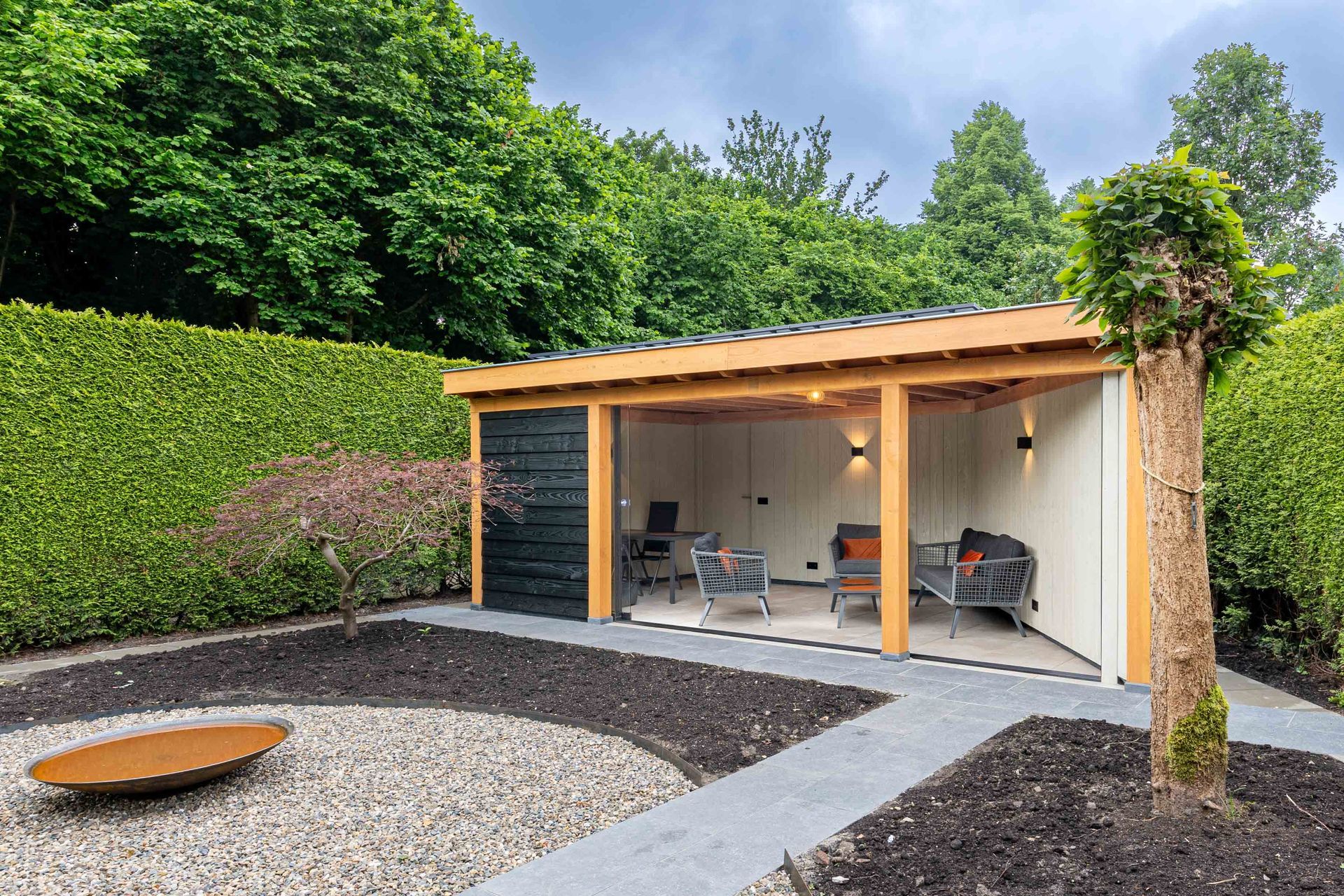 Een moderne houten tuinlounge met een open voorkant, zitgelegenheid buiten en een aangelegde grindtuin met een metalen vuurkorf.