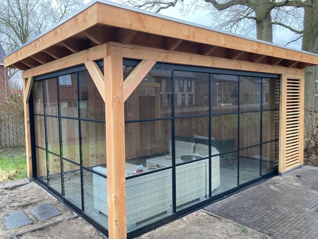 Een moderne houten tuinlounge met zwart omlijste glazen wanden en een zijpaneel met lamellen, geplaatst in een achtertuin.