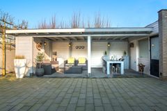 Een overdekt terras met een zithoek, eettafel en stoelen in een betegelde achtertuin, met een witte constructie en grijs meubilair.
