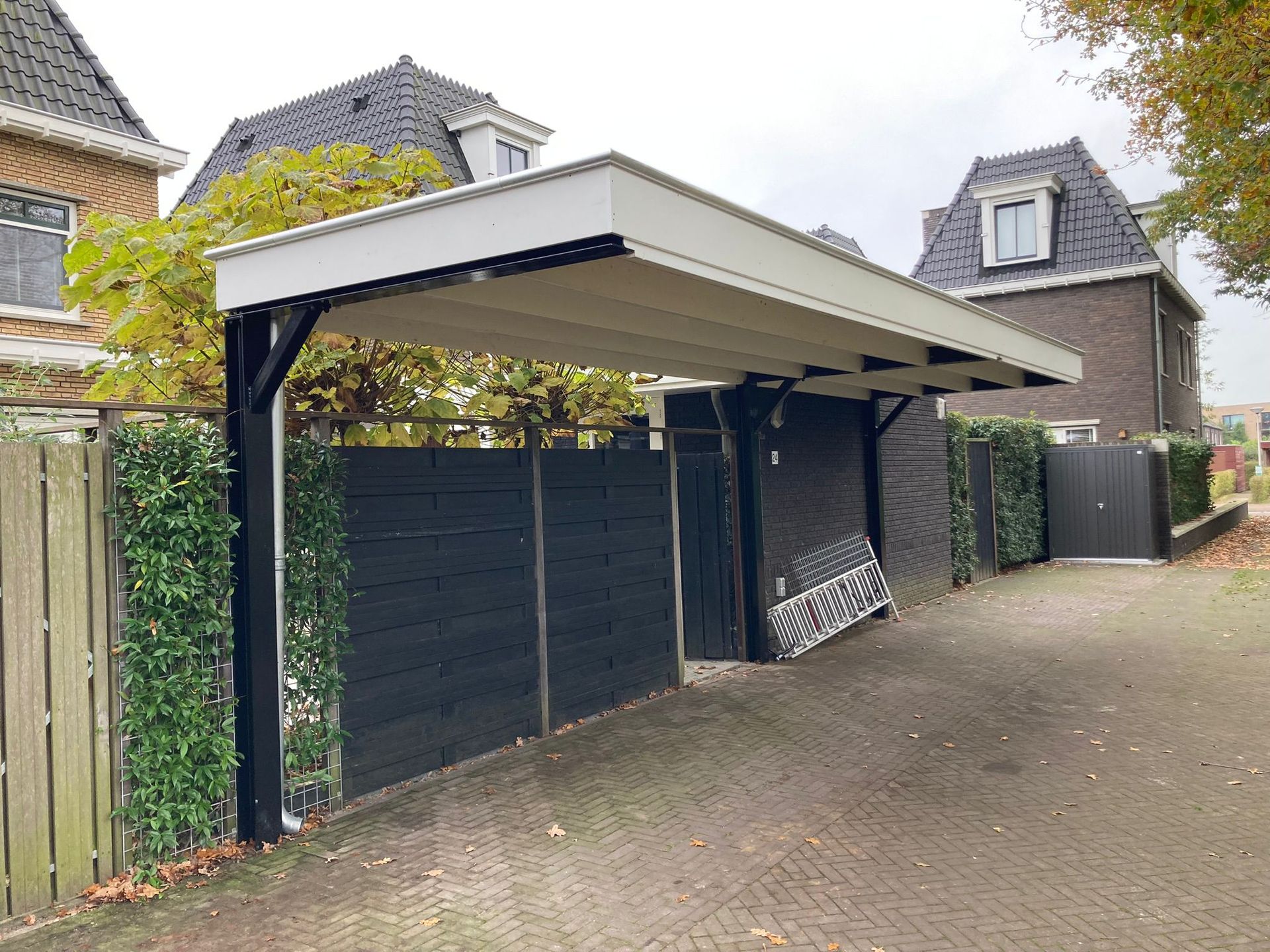 Een moderne carport met zwarte palen en een crèmekleurig dak staat naast een donkere houten schutting in een woonwijk.