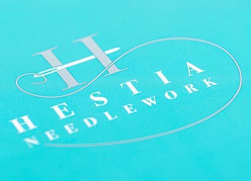 The logo for hestia needlework is on a blue background