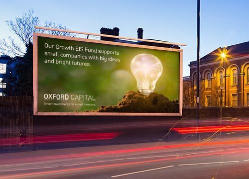 a large billboard with an advert for oxford capital shows a light bulb on top of it is on the side of the road .